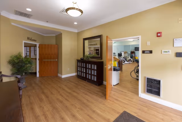 Interior view of a rehabilitation facility hallway with wooden flooring, beige walls, a wooden cabinet with a mirror above it, a plant, and an open door leading to a therapy services room with exercise equipment visible inside.