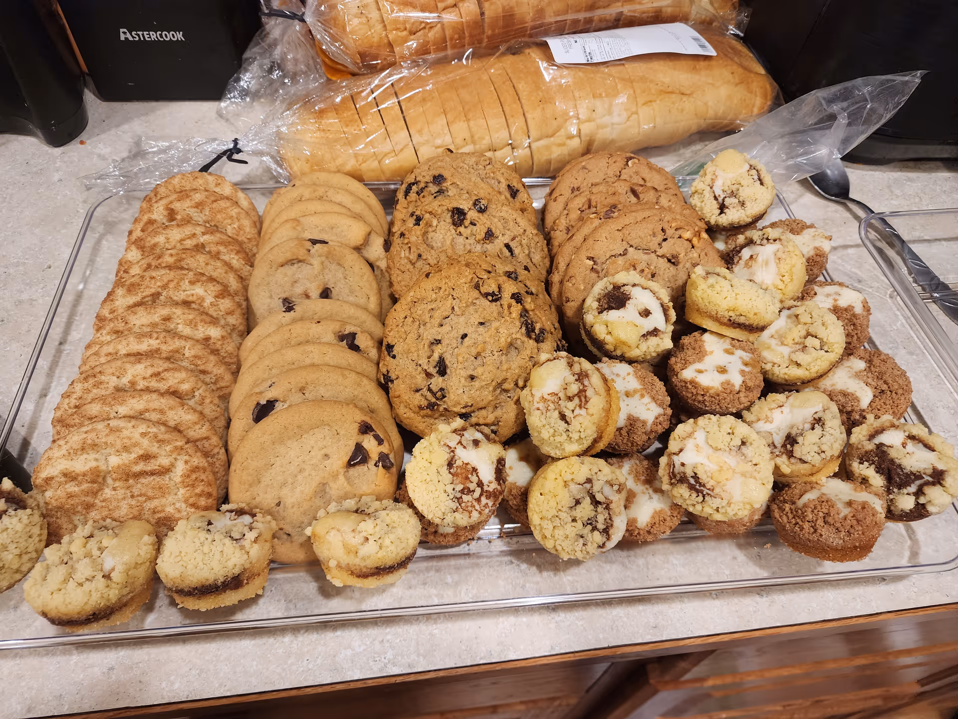 A clear rectangular tray filled with an assortment of baked goods including snickerdoodle cookies, chocolate chip cookies, oatmeal raisin cookies, and mini crumb-topped muffins with icing. Behind the tray, there are two loaves of sliced bread in plastic packaging on a kitchen countertop.