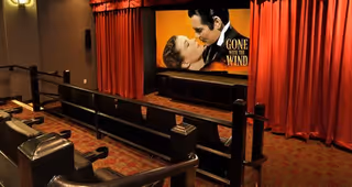 A small theater room with rows of comfortable seats facing a large screen displaying the movie poster for 'Gone with the Wind'. The room has red curtains on the sides of the screen and warm lighting.