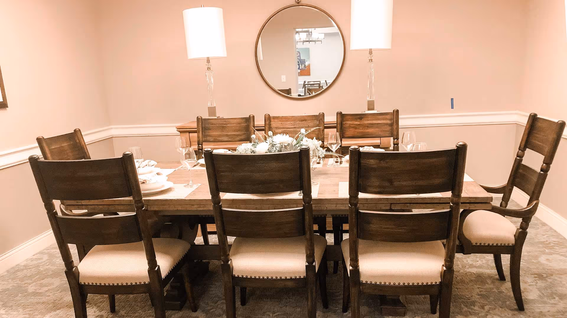 Wooden dining table with eight chairs, place settings and a floral centerpiece beneath two lamps and a round wall mirror in a beige dining room.