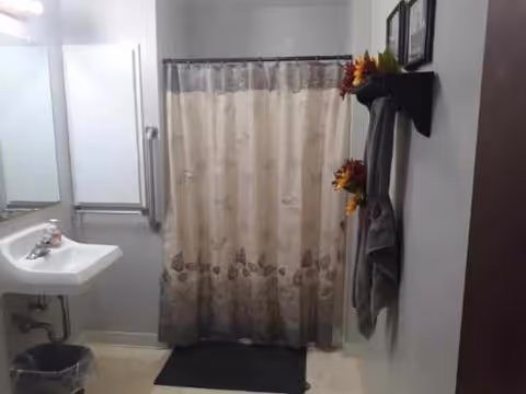 A bathroom with a white sink and mirror on the left, a shower with a beige and brown floral shower curtain in the center, a black floor mat in front of the shower, and a wall-mounted shelf on the right holding decorative flowers and a hanging gray towel.