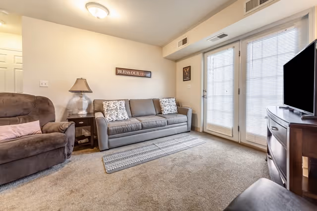 A cozy living room with a gray sofa adorned with patterned pillows, a brown recliner chair with a pink cushion, a wooden side table with a lamp, a TV on a wooden stand, and glass doors with blinds letting in natural light. A wall decoration reads 'BLESS OUR HOME'.