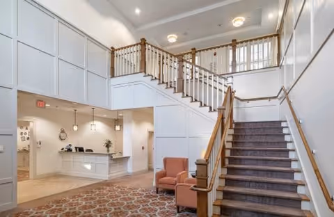 Bright reception lobby with a carpeted staircase, seating area, and front desk under high ceilings.