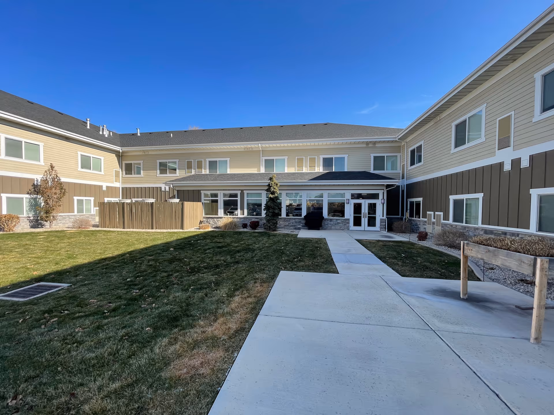Entrance courtyard and exterior facade of a two-story assisted living building with a sidewalk and lawn under a clear blue sky.