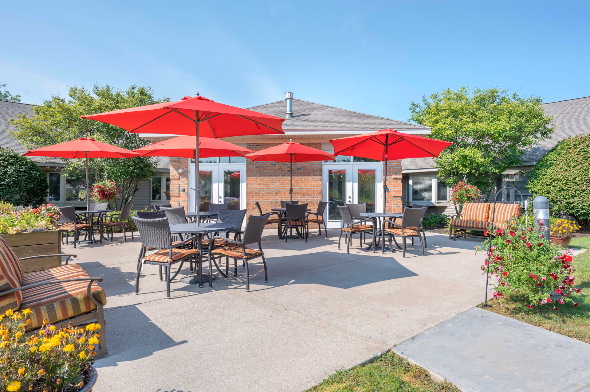 Outdoor patio area at Brookdale Granger with several tables and chairs under red umbrellas, surrounded by greenery and flowers, with a brick building in the background.