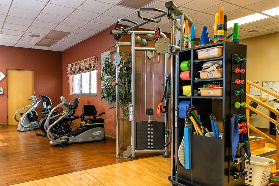 Interior view of a fitness room in a senior living facility with exercise equipment including stationary bikes, a multi-functional weight machine, and a storage rack holding various exercise accessories such as cones, resistance bands, and small weights. The room has wooden floors, a window with a valance, and a reddish-brown accent wall.