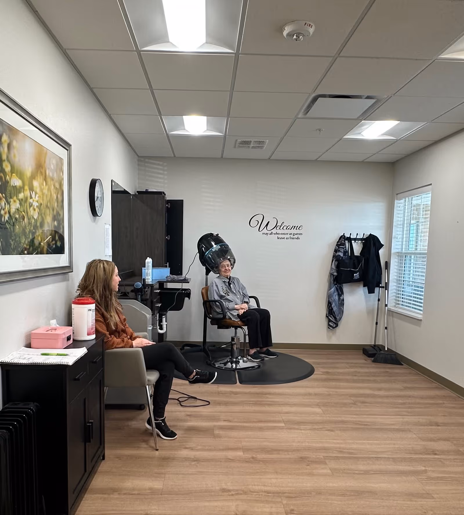 A senior woman sitting under a hair dryer in a salon area of a senior living community. Another woman is seated nearby, waiting. The room has light-colored walls, a wooden floor, a wall clock, a framed picture of flowers, and a coat rack with hanging items. A wall decal reads 'Welcome may all who enter as guests leave as friends.'