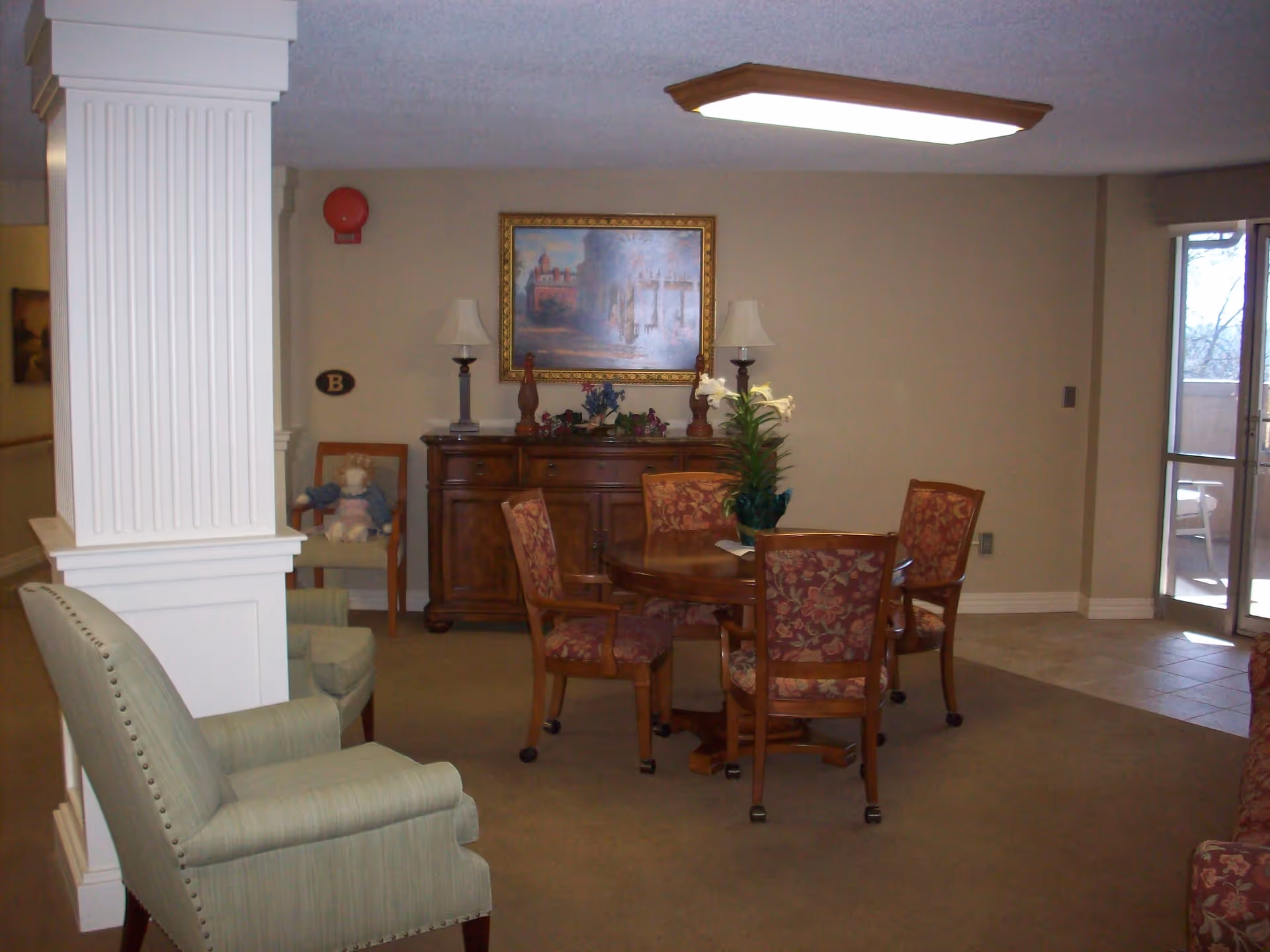 A cozy senior living common area with a round wooden table surrounded by four floral upholstered chairs on wheels. Behind the table is a wooden sideboard with two lamps and decorative items, including a framed painting on the wall above it. To the left, there is a green armchair and a chair with a doll sitting on it. The room has beige walls, carpeted floor, and a large window or glass door letting in natural light.