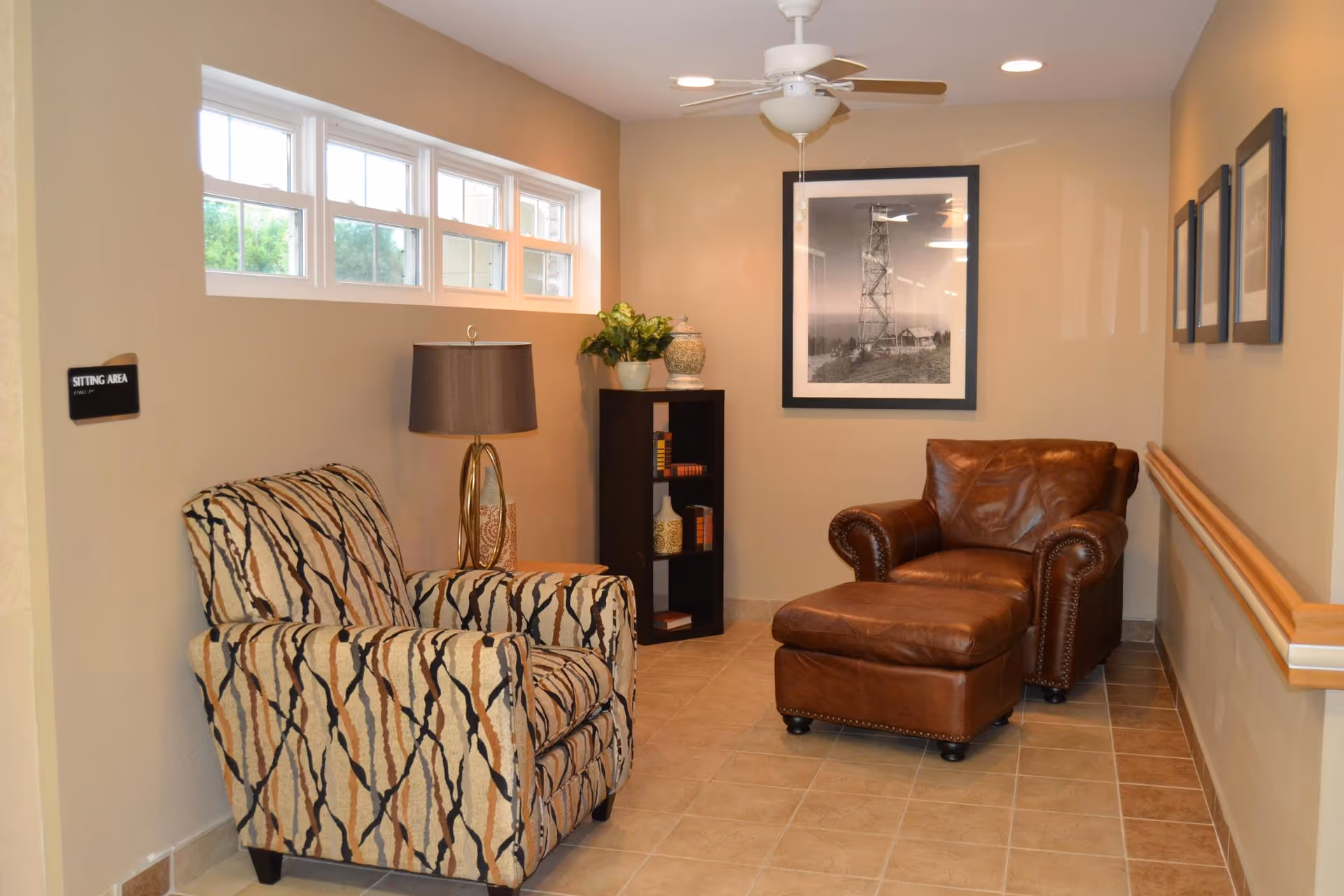 A cozy sitting area in a hallway with a patterned armchair, a brown leather armchair with matching ottoman, a floor lamp, a small black bookshelf with decorative items, and framed pictures on the walls. There is a small window near the ceiling letting in natural light.