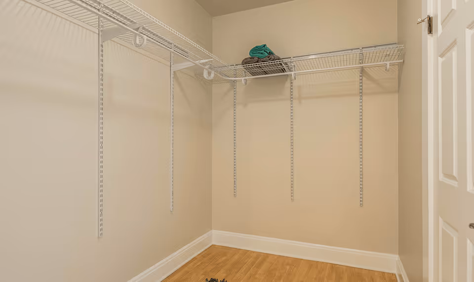 Small empty walk-in closet with white wire shelving, beige walls, wood floor, and a few folded towels on the top shelf.