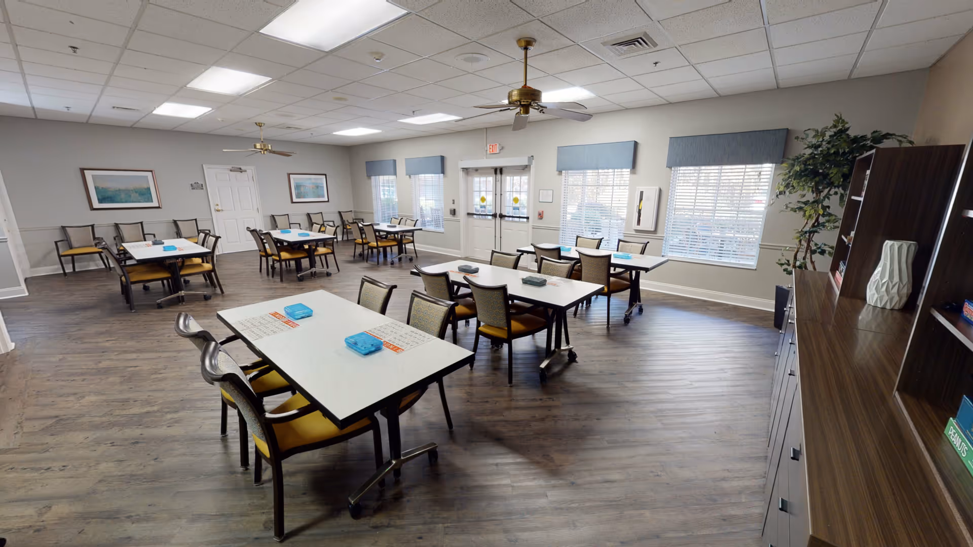 A spacious dining room with multiple tables and chairs arranged neatly. The room has large windows with blue valances, ceiling fans, and wood flooring. There are framed pictures on the walls and a cabinet with decorative items on the right side.