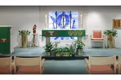 Small chapel interior with an altar draped in green cloth, a blue stained-glass window behind it, rows of chairs and potted plants
