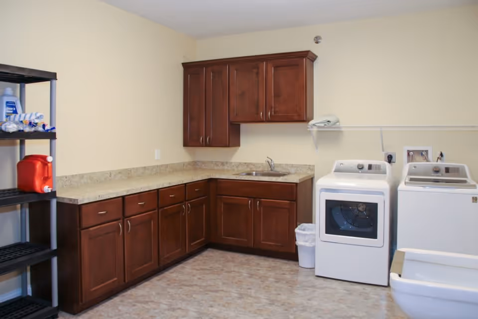 Laundry room with dark wood cabinets, a countertop with a sink, a white washing machine and dryer, a small trash can, and a black shelving unit holding laundry supplies.