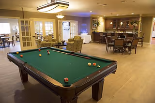 Spacious senior living common room with a pool table in the foreground, seating, dining tables, and a bar in the background.
