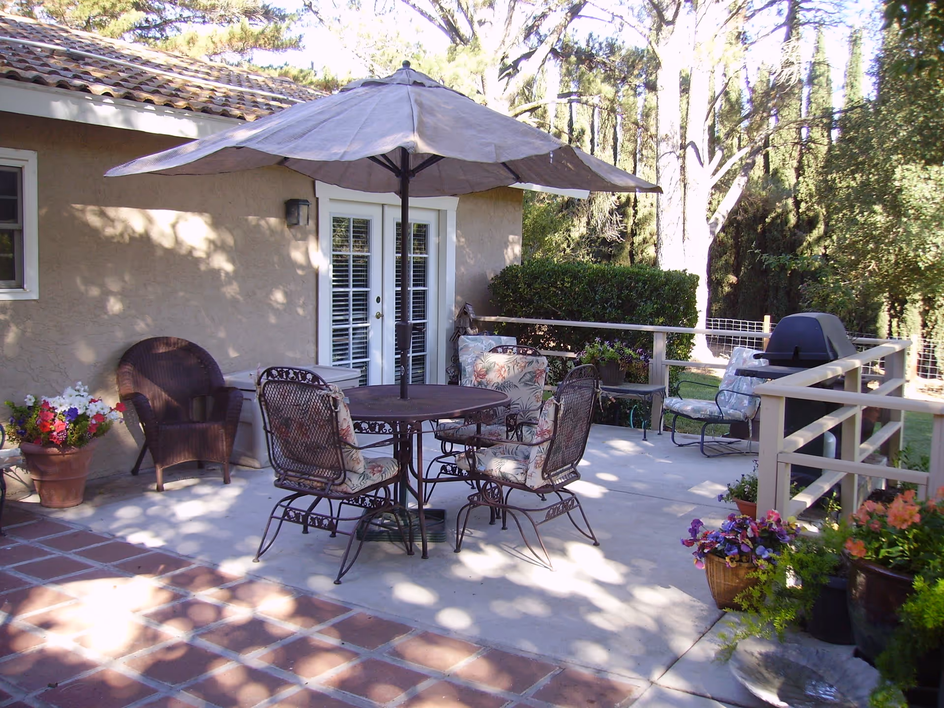 Outdoor patio area with a round table and four cushioned metal chairs under a large umbrella. There are additional chairs, potted plants with colorful flowers, a barbecue grill, and a beige building with French doors in the background. Trees and greenery surround the patio.