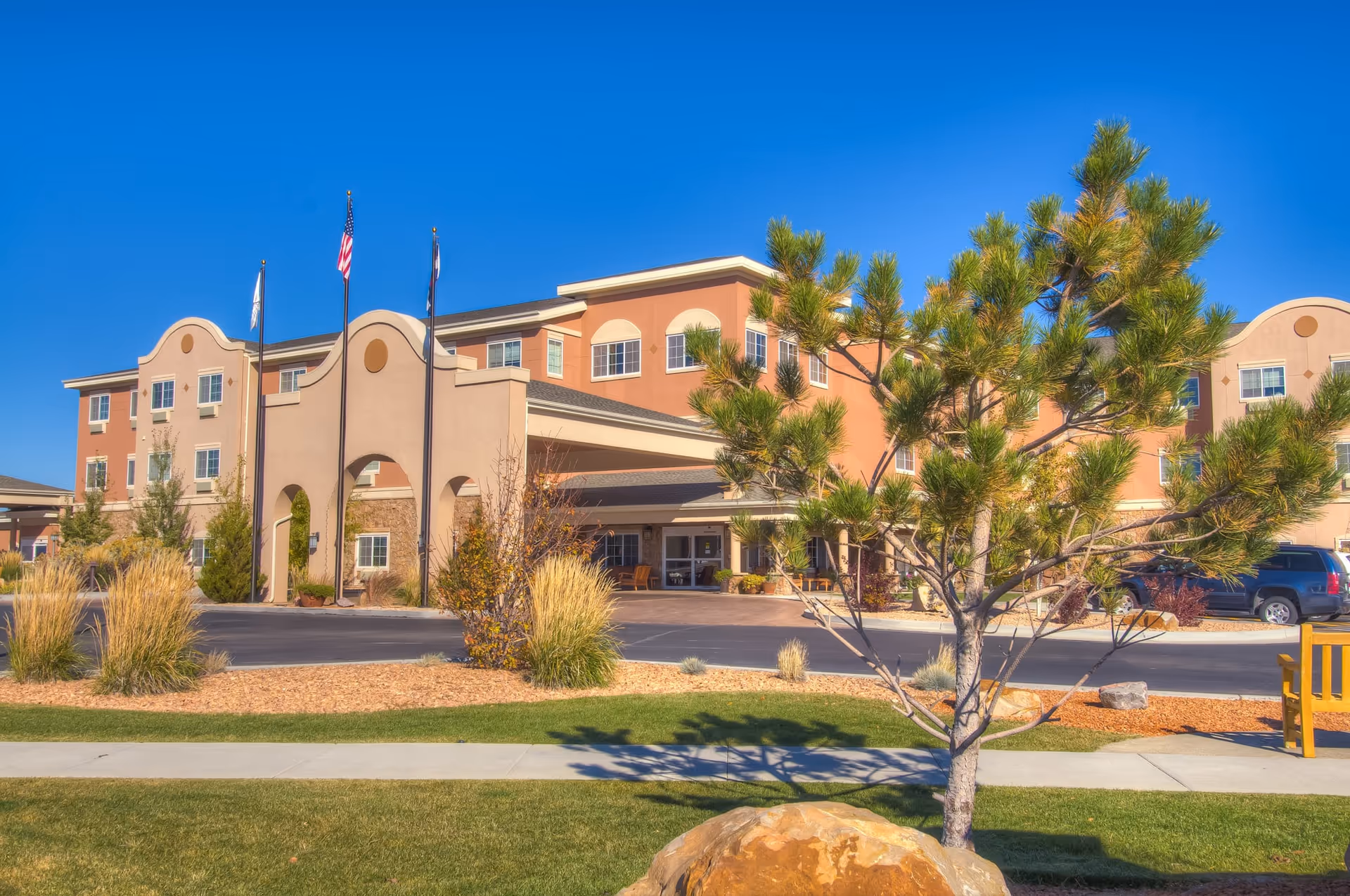 Exterior view of a senior living facility named The Bridge at Alamosa, showing a three-story building with a covered entrance, three flagpoles with flags, landscaped grounds with grass, bushes, a tree, and a clear blue sky.