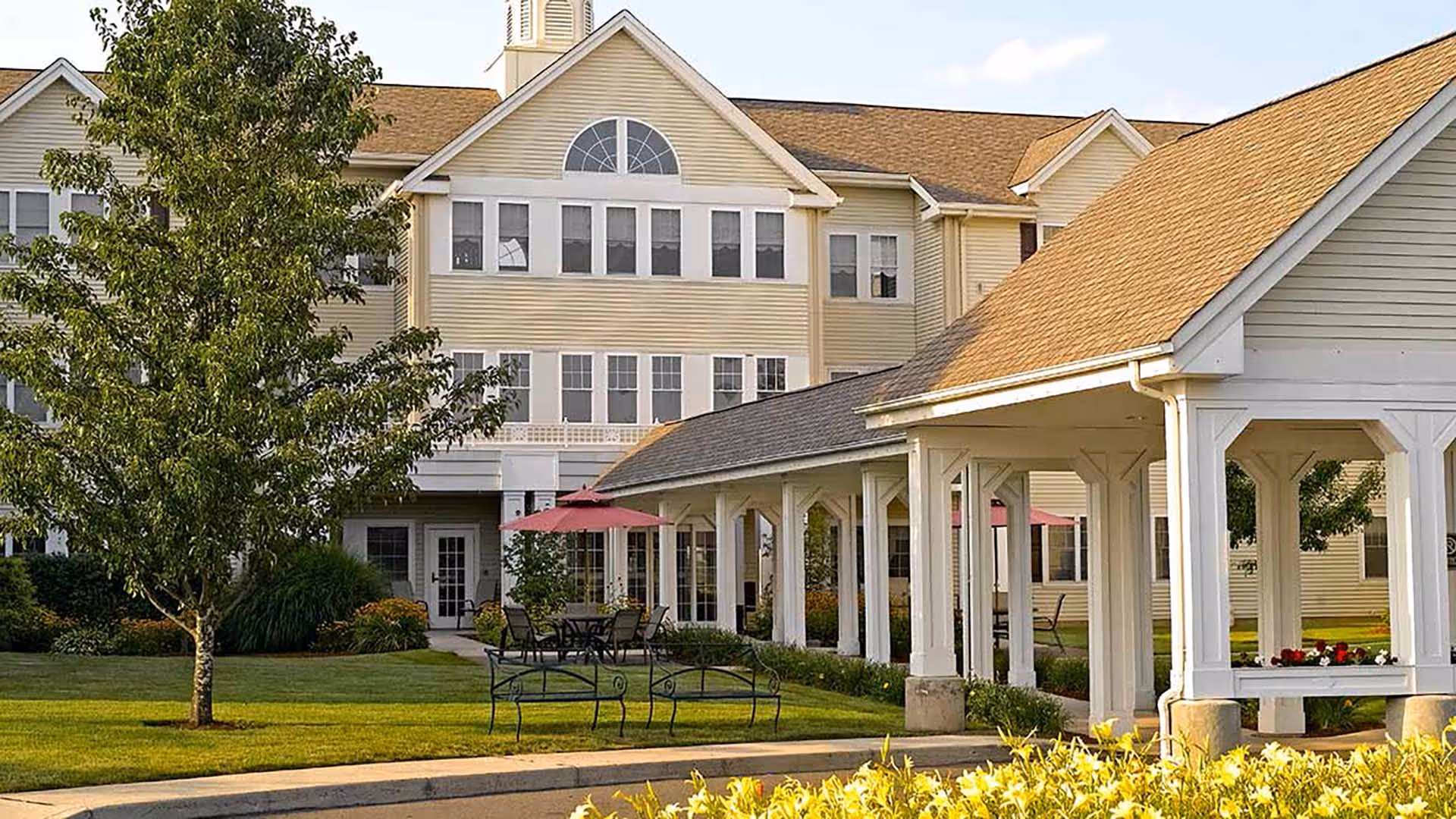 Exterior view of a senior living facility building with beige siding and multiple windows. There is a covered walkway supported by white columns, a green lawn with a tree, benches, and outdoor tables with red umbrellas.