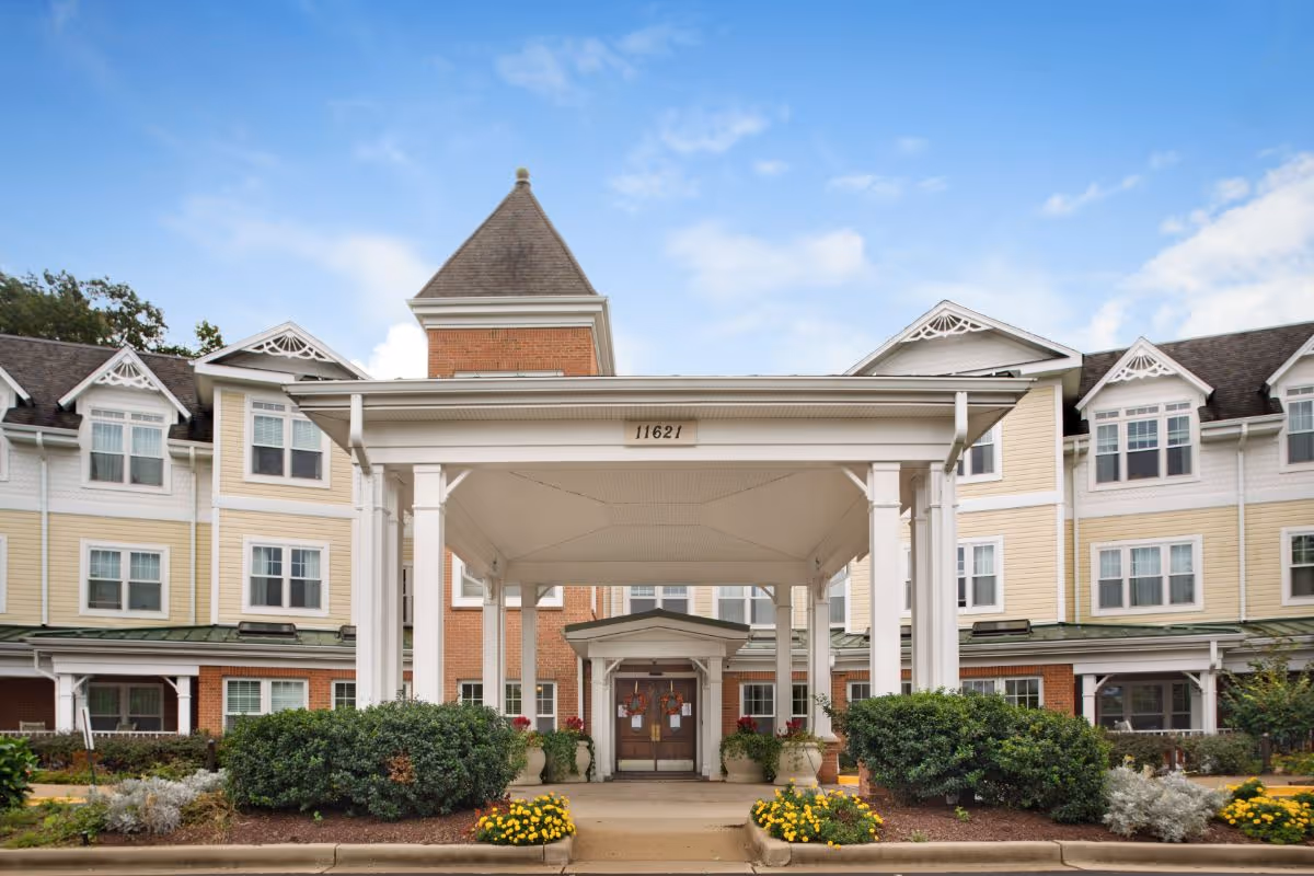 Exterior front entrance of a three-story senior living building with a covered porte-cochère, landscaping, and the address "11621" above the canopy.