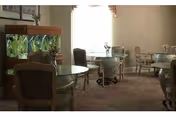A softly lit room with several round tables and cushioned chairs arranged around them. There is a large fish tank on the left side of the room, and windows with sheer curtains allowing natural light to enter. The room has a carpeted floor and a calm, inviting atmosphere.