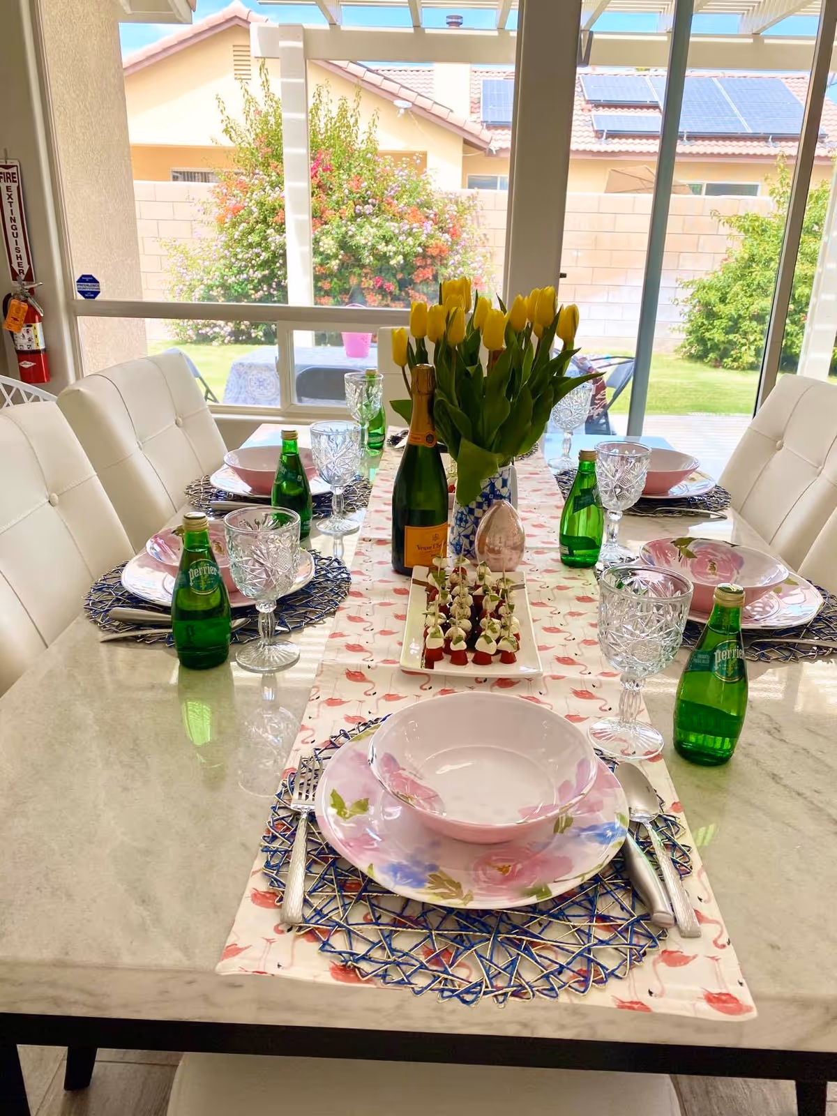 A dining table set for six with white cushioned chairs. The table has a floral table runner, pink floral plates and bowls, crystal wine glasses, green Perrier bottles, and a centerpiece of yellow tulips in a blue and white vase. Outside the large windows, there is a garden with blooming bushes and a patio area.