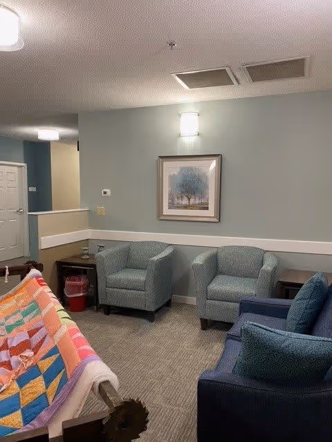 A cozy sitting area in a senior living facility with three upholstered armchairs arranged around a small table. A colorful quilt is draped over a wooden rack in the foreground. The walls are painted light green with a white chair rail, and a framed picture of trees hangs on the wall. The room is softly lit by ceiling and wall lights.