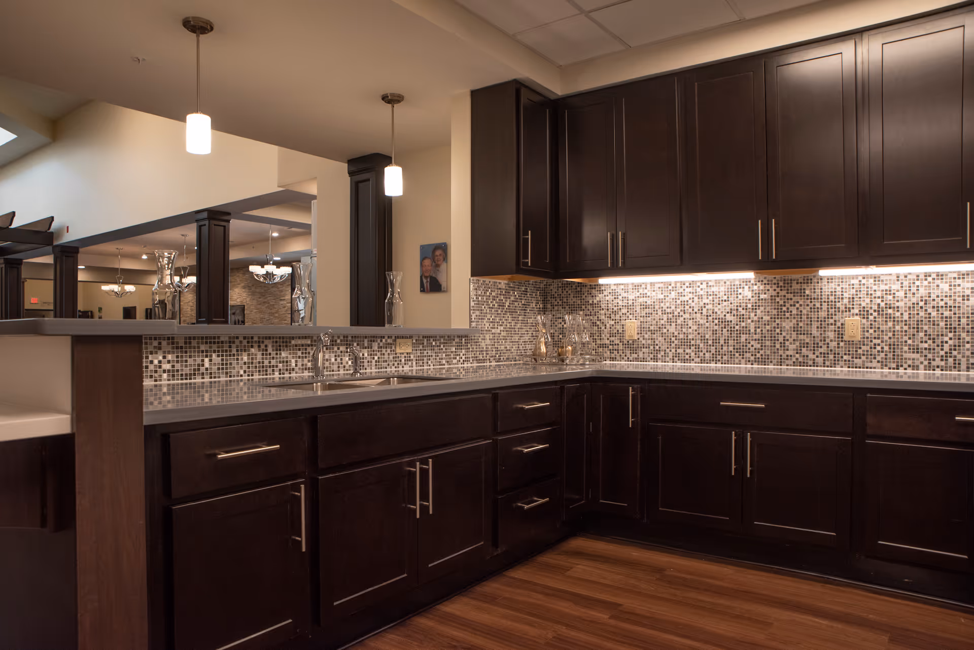 Modern kitchen area with dark wood cabinets, a mosaic tile backsplash, under-cabinet lighting, and a double sink. The kitchen opens up to a larger room with visible columns and pendant lights hanging from the ceiling.
