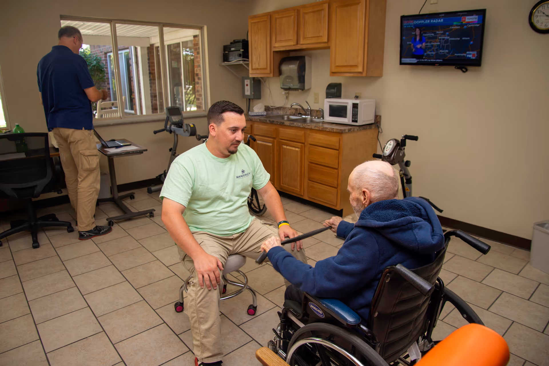 A man in a light green Manderley Health Care Center shirt is seated on a stool, engaging with an elderly man in a wheelchair inside a room with tiled floors and wooden cabinets. Another man is standing near a window working on a laptop. A television is mounted on the wall showing a weather forecast.