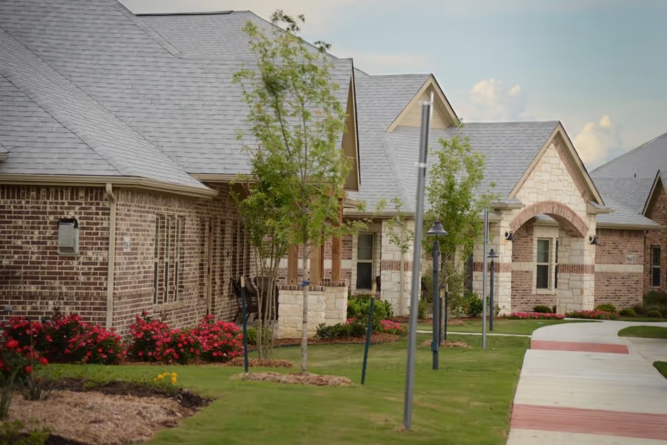 Exterior view of a senior living facility with brick and stone buildings, a well-maintained lawn, young trees, flower beds with red flowers, and a paved walkway with street lamps.