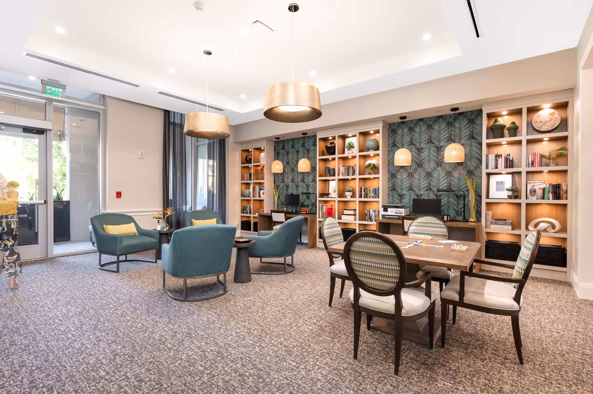 A bright and spacious common area in a senior living facility featuring a seating arrangement with four teal armchairs around a small round table, and a wooden table with four chairs nearby. The back wall has built-in wooden shelves filled with books, plants, and decorative items, with a green leafy patterned wallpaper behind them. Two large pendant lights hang from the ceiling, and there is a glass door leading outside on the left side. A person is partially visible walking out the door.