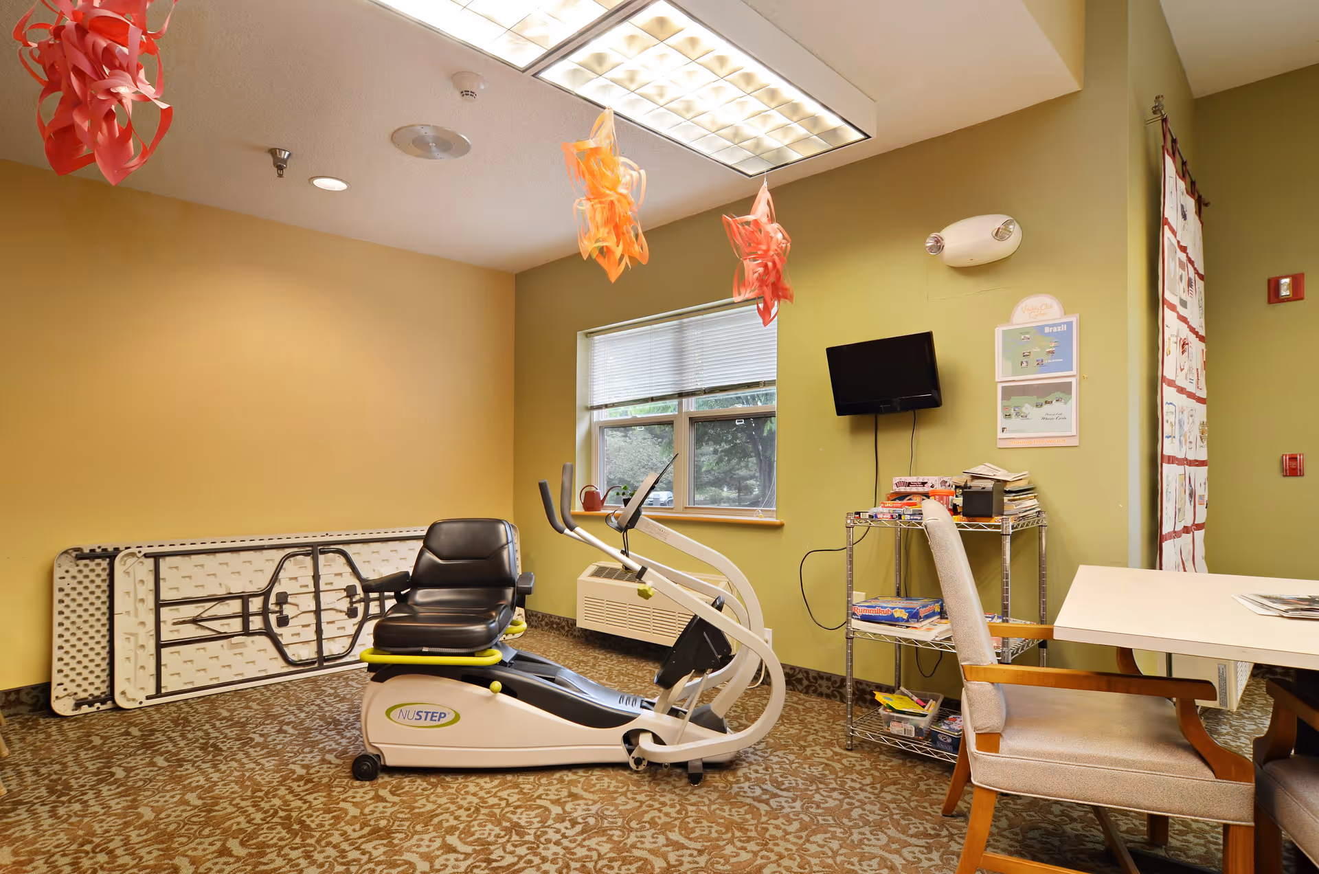A small exercise room with a recumbent exercise bike, a folding table leaning against the wall, a window with blinds, a small metal shelf holding board games and books, a wall-mounted TV, and a table with chairs. The walls are painted yellow and green, and there are decorative hanging paper ornaments from the ceiling.