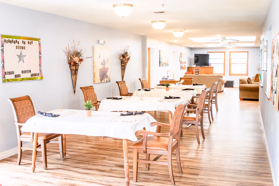 A bright and spacious dining area in a senior living facility with several tables covered in white tablecloths, each set with napkins and small potted plants. Wooden chairs with cushioned seats surround the tables. The room has light-colored walls decorated with floral artwork and wall-mounted flower arrangements. In the background, there is a living area with sofas, a TV, and large windows letting in natural light.