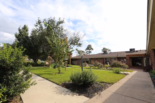 Outdoor courtyard area at Timberwood Nursing & Rehabilitation Center featuring a green lawn, small trees, shrubs, flower beds, and paved walkways surrounded by single-story brick buildings under a partly cloudy sky.