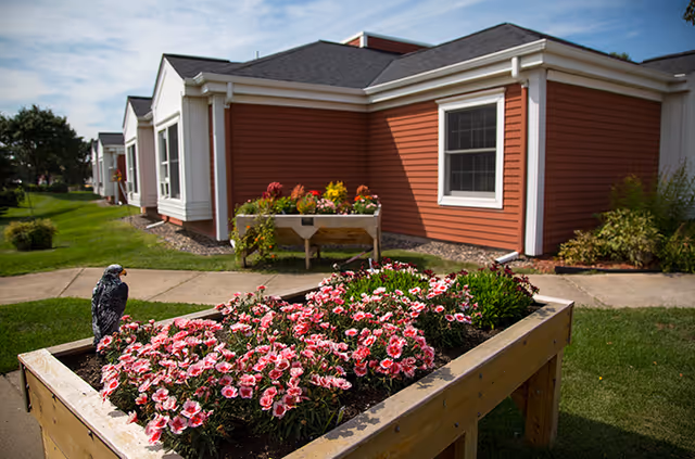 Raised wooden planter filled with pink flowers in front of a red single-story senior living building and a walkway under a partly cloudy sky.
