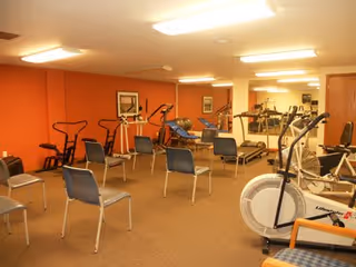 Exercise/fitness room with stationary bikes, various exercise machines and rows of chairs against orange walls.