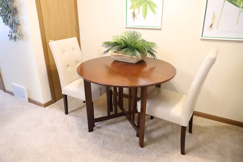 A small round wooden dining table with a potted green plant centerpiece, accompanied by two white upholstered chairs in a carpeted room with light-colored walls and botanical artwork hanging on the wall.