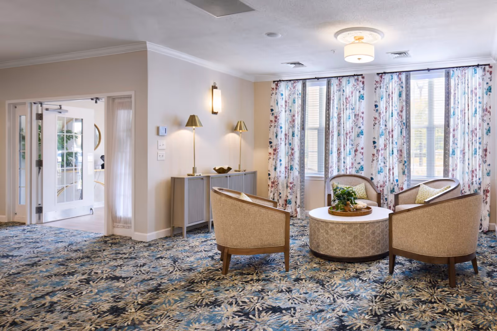 A cozy sitting area in a senior living facility with four beige armchairs arranged around a round coffee table. The room features large windows with floral curtains, a patterned carpet, and soft lighting from wall sconces and a ceiling light. A sideboard with two lamps is placed against the wall near a set of double glass doors.