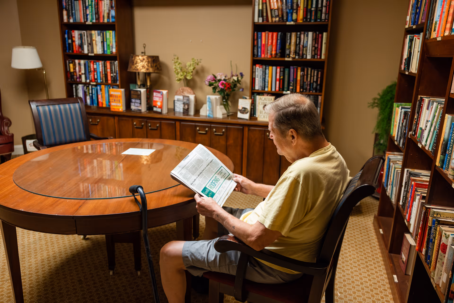 An elderly man sitting in a chair reading a newspaper in a cozy library room with wooden bookshelves filled with books, a round wooden table with a glass top, and decorative items including flowers and a lamp.