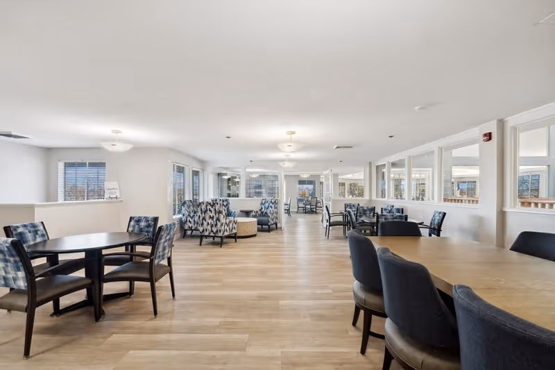 A spacious, well-lit common area with multiple seating arrangements including round tables with chairs and patterned armchairs. The room has large windows allowing natural light to fill the space, light-colored walls, and wooden flooring. Ceiling lights provide additional illumination.