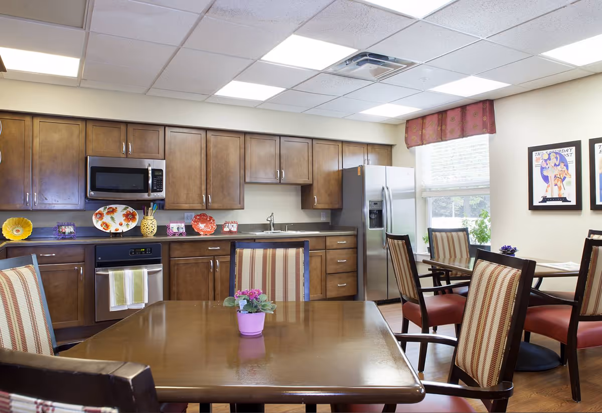A bright kitchen and dining area with wooden cabinets, stainless steel appliances including a microwave, oven, and refrigerator. There are several tables with striped cushioned chairs, a small potted plant on the table in the foreground, and framed artwork on the wall near a window with a red valance.