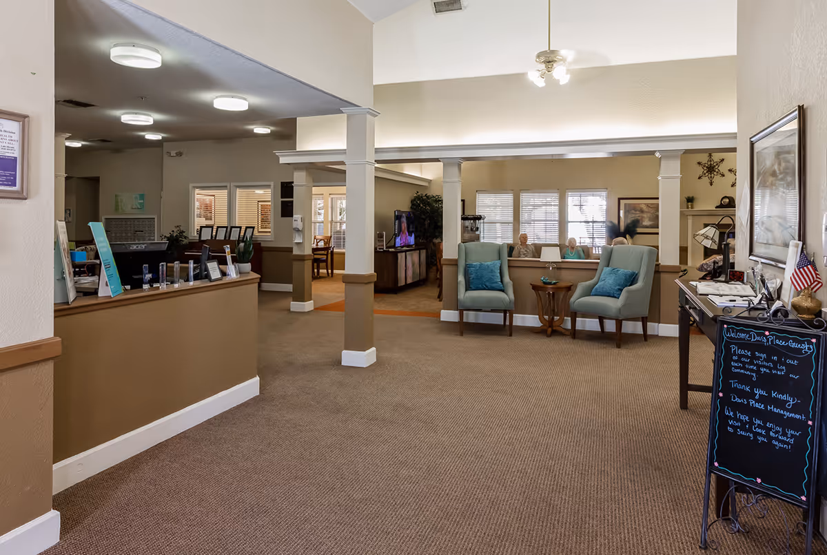 Interior view of a senior living facility reception area with a front desk on the left, two armchairs with blue cushions and a small table in the center, and a blackboard sign on the right welcoming guests. There are windows and a TV in the background, along with some seated residents and a person walking.