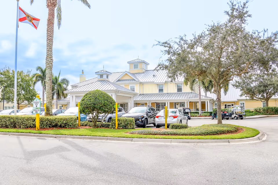 Exterior view of The Brennity at Melbourne Senior Living facility showing a light yellow building with a metal roof, surrounded by trees, bushes, and parked cars. A Florida state flag is flying on a flagpole near the entrance.