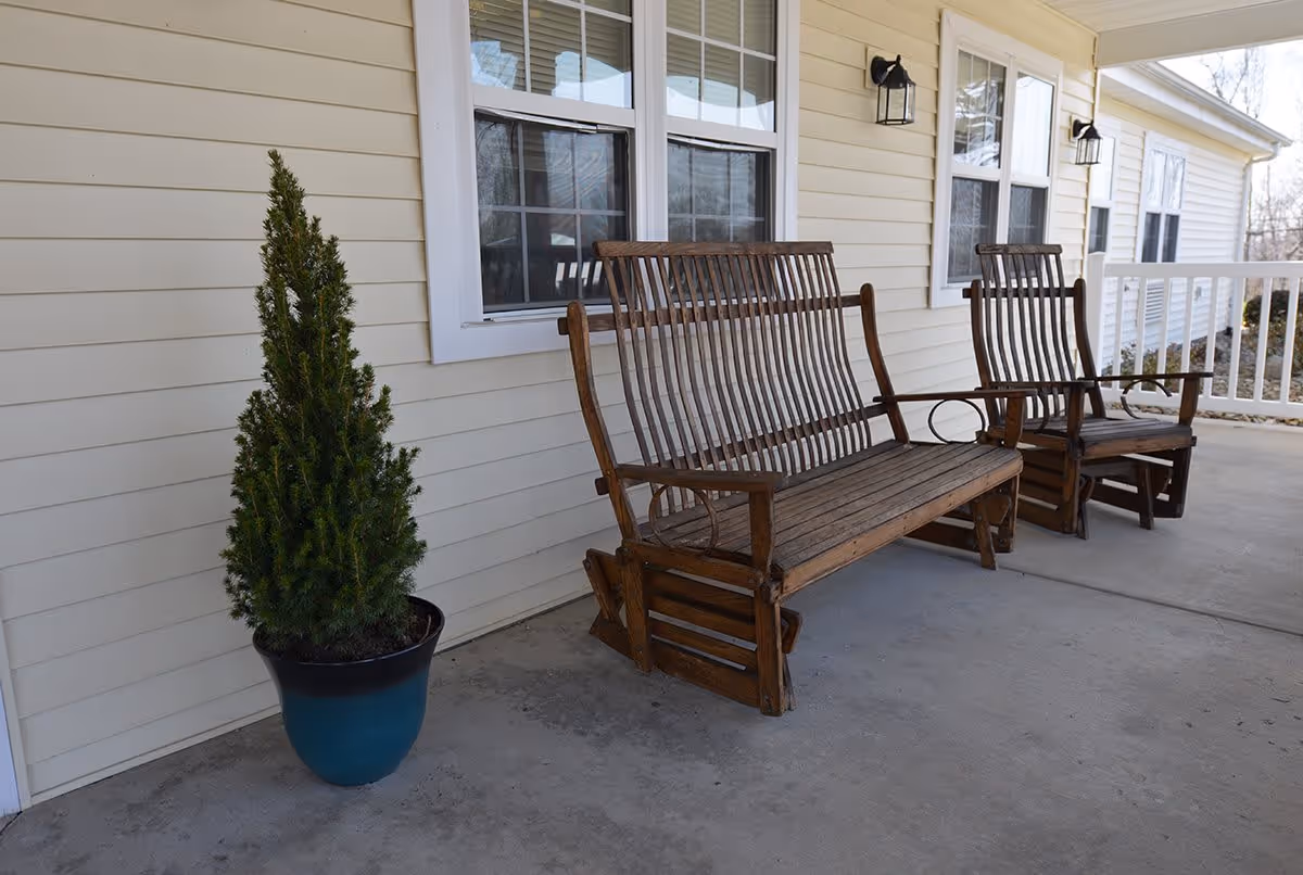 A porch area with two wooden rocking chairs and a wooden bench placed against the exterior wall of a light yellow building. There is a potted evergreen plant on the left side, and two windows with white frames above the seating. Two black wall-mounted lantern lights are visible on the wall.