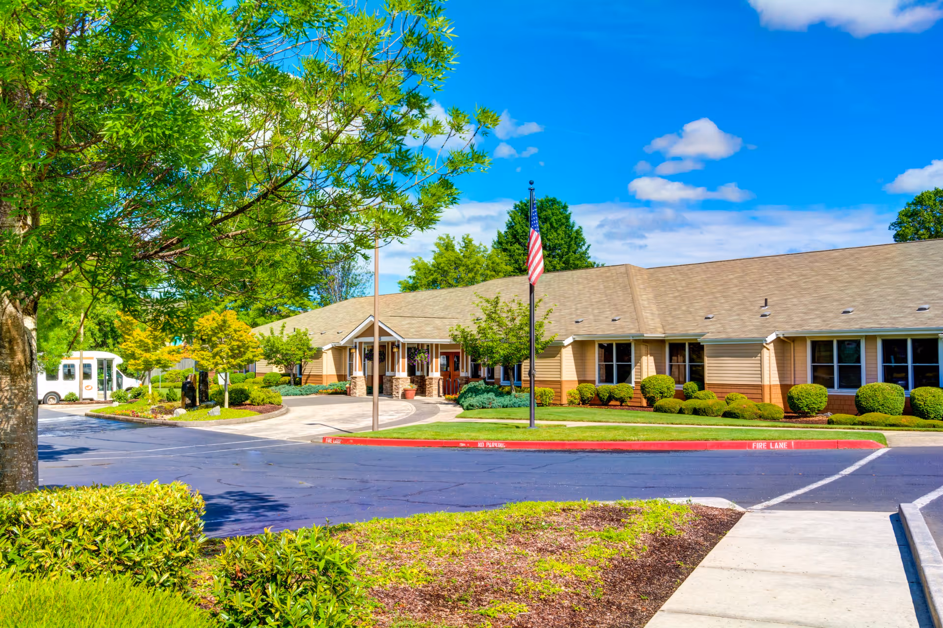 Exterior view of Monterey Court Memory Care facility showing a single-story building with beige siding and a tan roof, surrounded by well-maintained landscaping including green bushes, trees, and a lawn. There is a driveway and parking area in front with a fire lane marked in red, and an American flag on a flagpole near the entrance under a clear blue sky with some clouds.