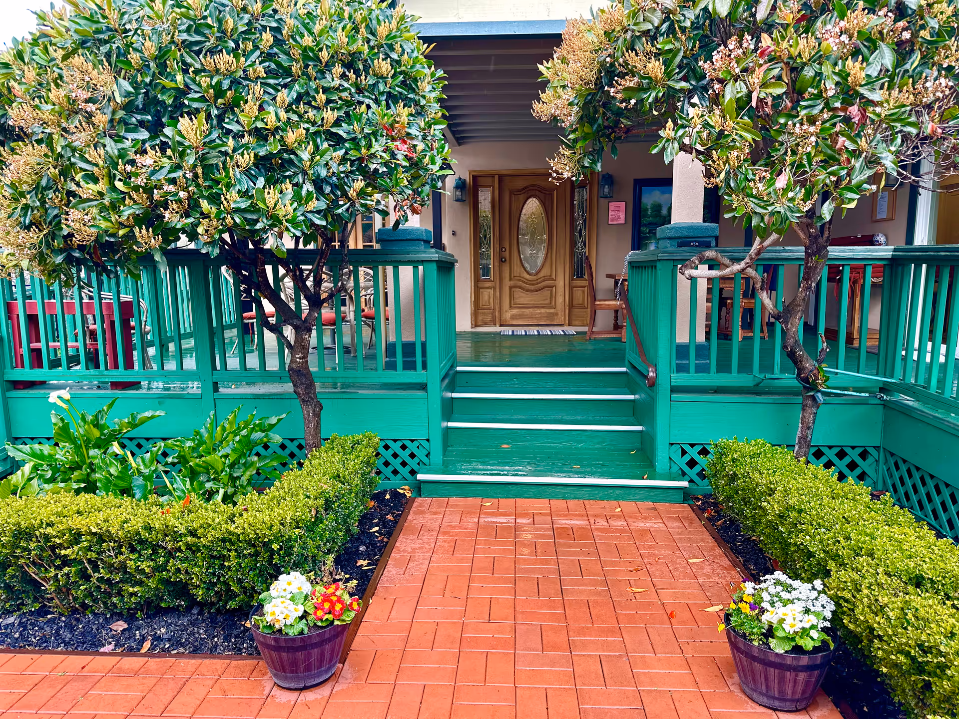 Front entrance of Bella Vista Village II featuring a green wooden porch with steps leading up to a wooden door with an oval glass panel. The porch is flanked by two small trees and neatly trimmed bushes, with flower pots containing colorful flowers on either side of the brick pathway.