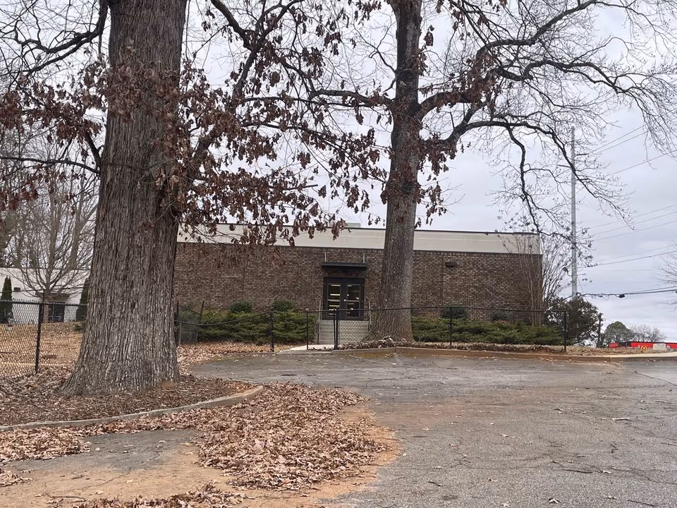 Brick single-story building entrance seen behind two large leafless trees with a fenced parking area and fallen leaves in the foreground.