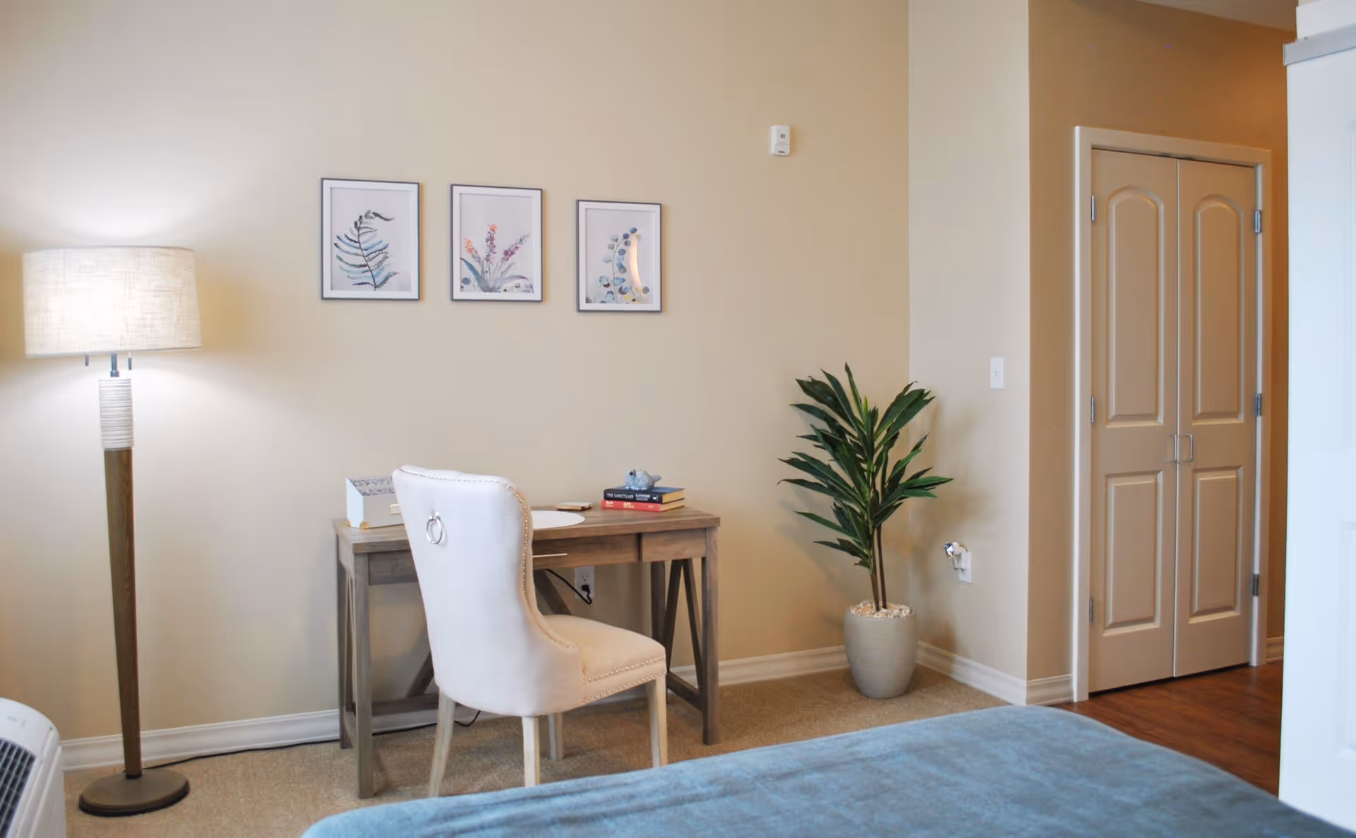 Bedroom nook with a wooden desk, upholstered chair, floor lamp, potted plant, wall art, and a partially visible bed.