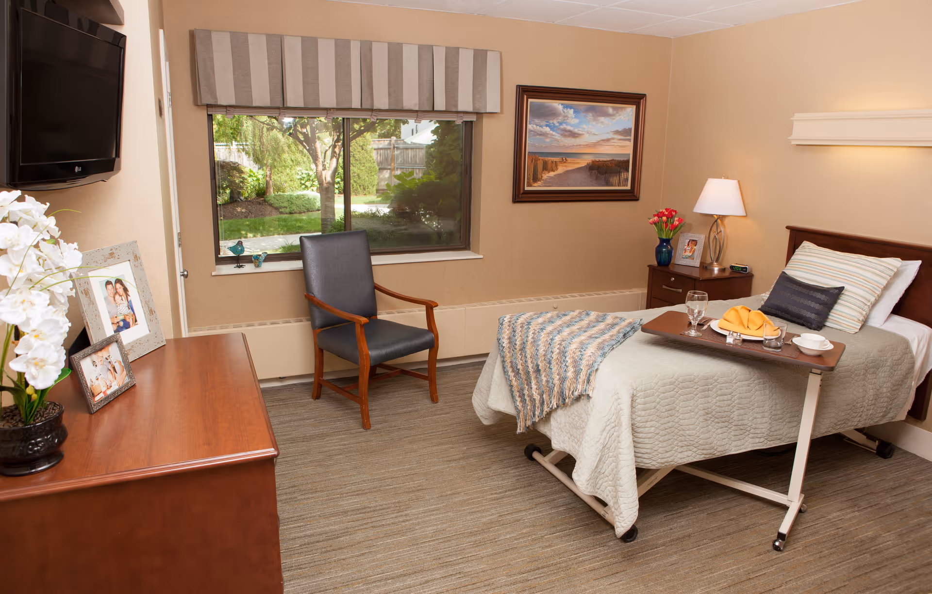 A cozy bedroom in a senior living facility with a single bed covered in a light gray quilt and a striped throw blanket. A wooden bedside table holds a lamp, a vase with red flowers, and a framed photo. A wooden chair with dark cushions is placed near a large window with a striped valance, showing a view of a garden outside. A wooden dresser with framed photos and a potted white orchid is on the left side of the room. A flat-screen TV is mounted on the wall above the dresser.