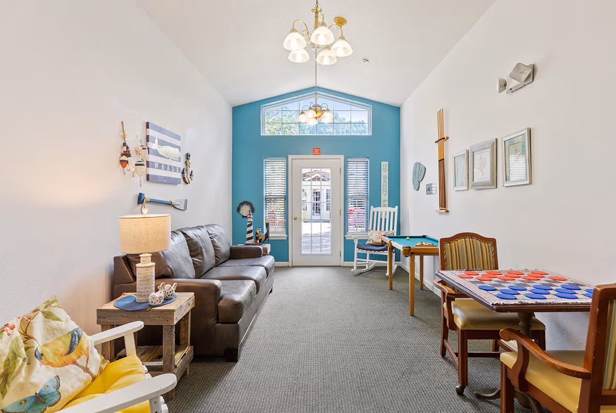 A bright and cozy common area with a brown leather sofa, a wooden side table with a lamp, a rocking chair with a colorful cushion, a small pool table, and a table with a checkers game set up. The room has white walls with some nautical-themed decorations and a blue accent wall with a glass door and windows letting in natural light.