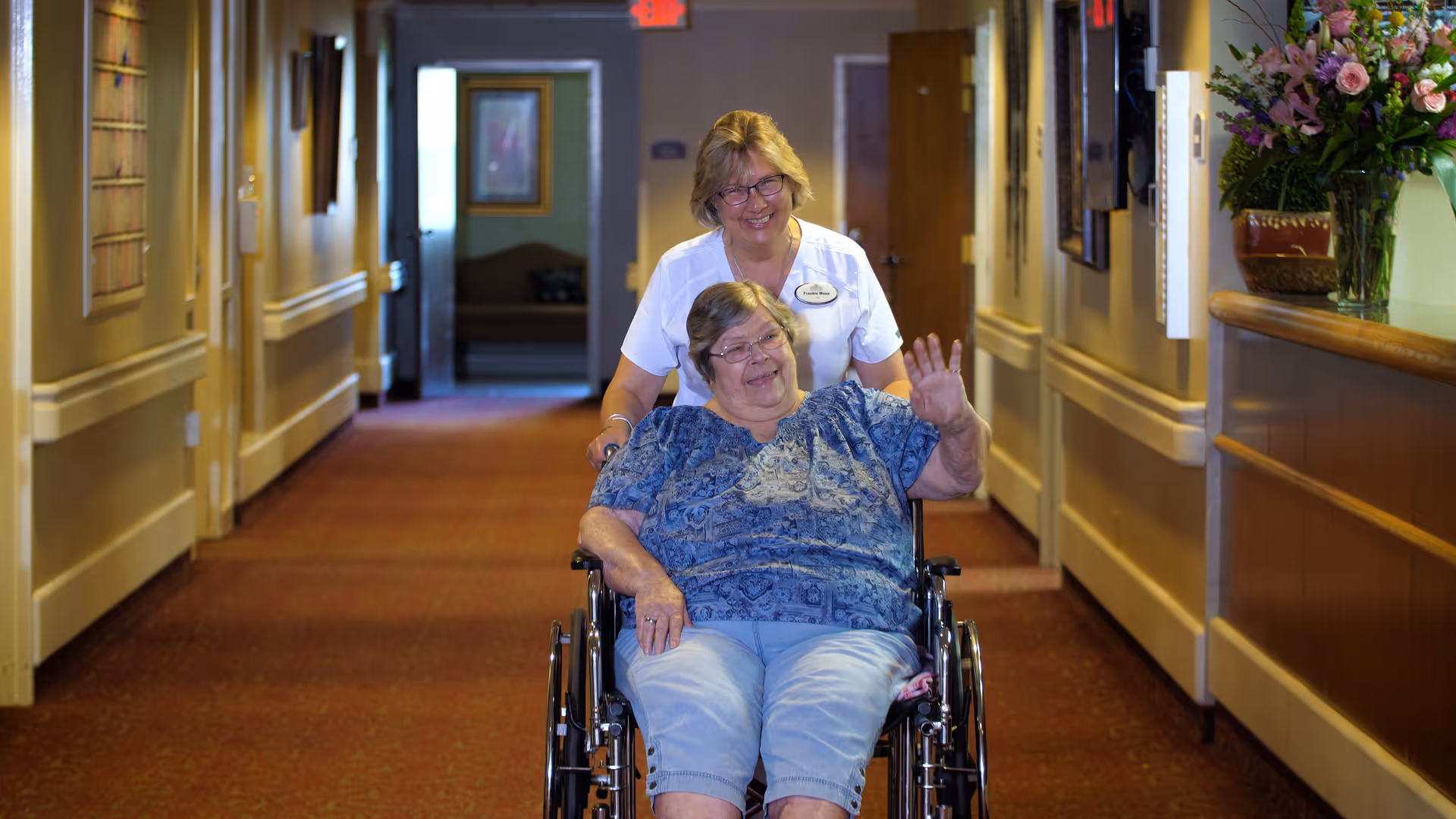 A smiling elderly woman in a wheelchair waving while being pushed down a hallway by a smiling female caregiver wearing a white uniform. The hallway has warm lighting, framed pictures on the walls, and a flower arrangement on a counter to the right.