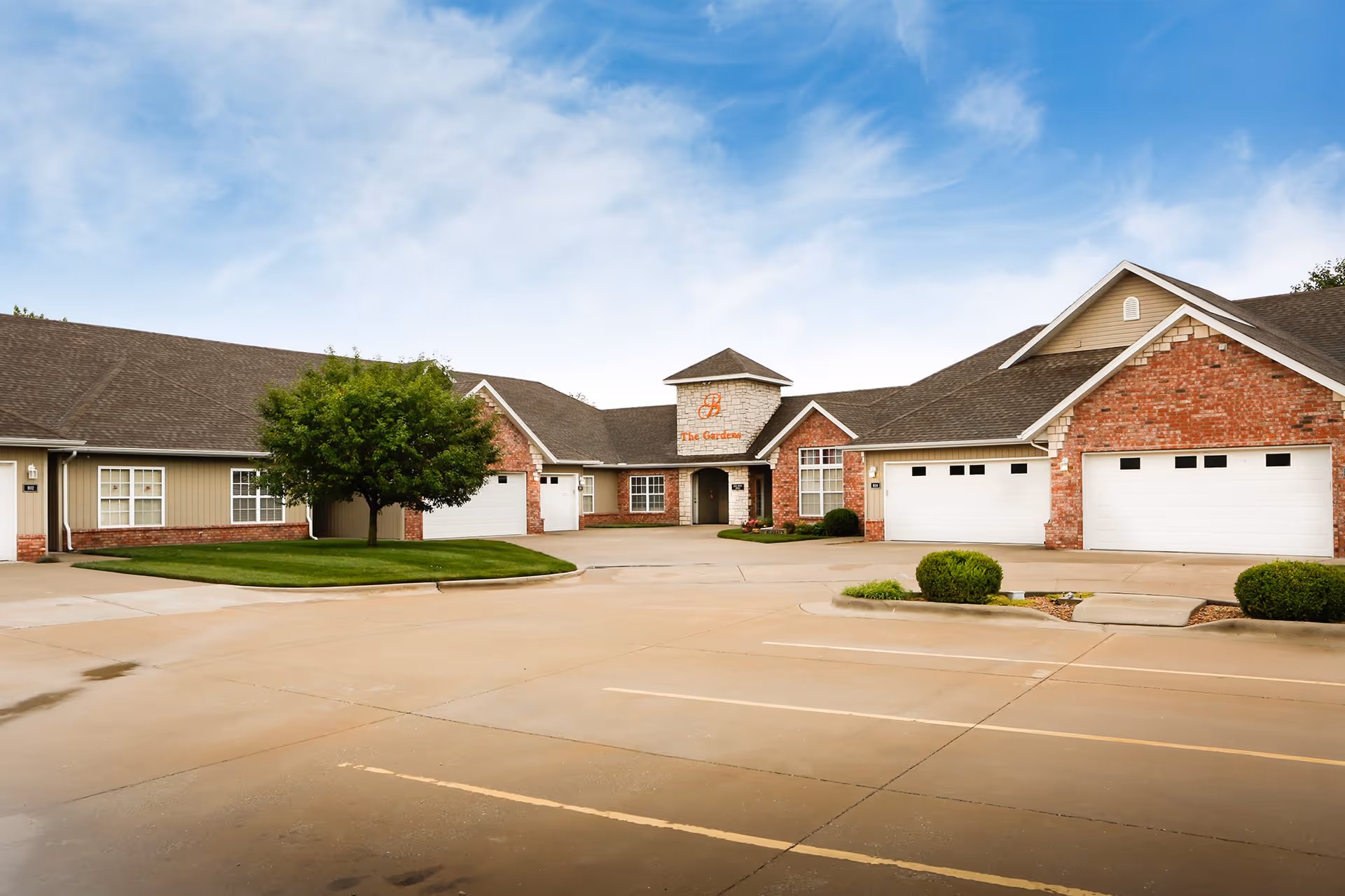 Front exterior of The Gardens independent living building with brick and siding, attached garages, a parking lot, and a small landscaped island under a blue sky.